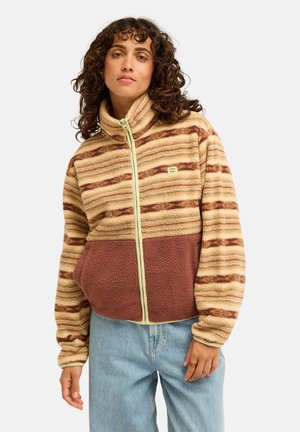 Femme aux cheveux bouclés portant une veste polaire zippée beige et marron rayée et un jean bleu clair sur un fond blanc uni.