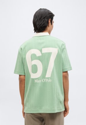 Homme aux cheveux courts et foncés portant une chemise à manches courtes vert clair avec les grands chiffres blancs "67" et le texte "Marc O'Polo" dans le dos.