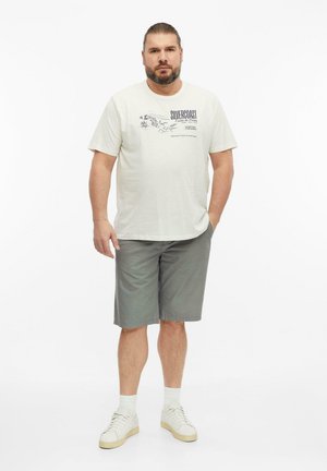 Mann mit Bart, der ein offwhite bedrucktes T-Shirt, graue knielange Shorts, weiße Socken und weiße Sneakers trägt und vor weißem Hintergrund steht.