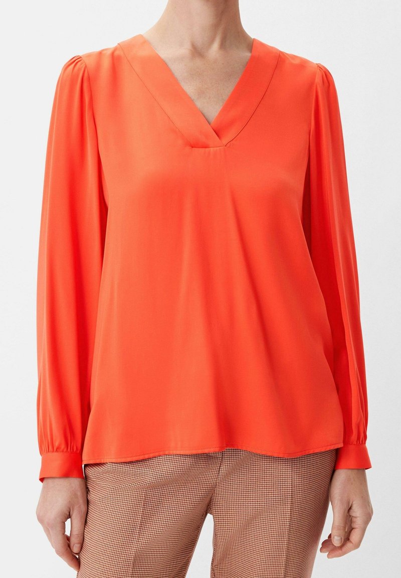 Blouse orange à col en V avec des manches longues, présentant un détail froncé à l'épaule et une texture lisse et brillante. Associée à un pantalon à motif pied-de-poule.