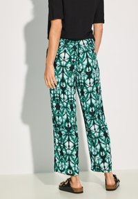 Väljande byxor i ett grönt och svart geometriskt mönster, med ett mjukt tyg och vida ben, tillsammans med mörka slide-sandaler.