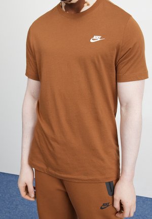 Homme portant un t-shirt et un short Nike marron, debout devant un fond blanc et bleu, avec les logos Nike visibles sur les deux vêtements.