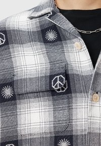 Chemise à carreaux noir et blanc avec un symbole de paix brodé et des motifs de soleil, dotée de boutons en bois et d'un tissu texturé.