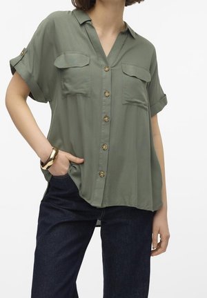 Donna che indossa una camicia ampia verde oliva a maniche corte con tasche sul petto e jeans blu scuro, con una mano in tasca e un braccialetto d'oro.