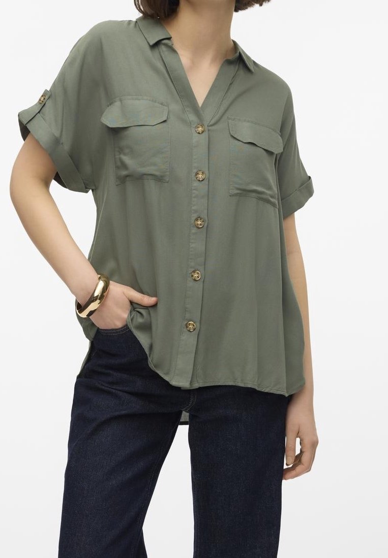 Femme portant une chemise ample à manches courtes vert olive avec des poches sur la poitrine et un jean bleu foncé, avec une main dans la poche et un bracelet en or.
