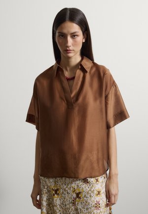 Mujer joven con cabello largo y oscuro, vestida con una camisa marrón de manga corta y una falda estampada, posando frente a un fondo claro y sencillo.
