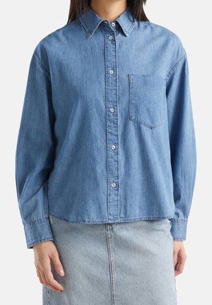 Femme portant une chemise en jean bleue boutonnée avec une poche poitrine et une jupe en jean bleu clair, debout devant un fond blanc.