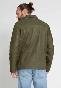 Grüne Wildlederjacke mit klassischem Kragen, nahtlosen Paneelen und taillierter Silhouette, kombiniert mit hellblauen Jeans.