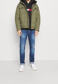 Giacca puffer verde oliva con cappuccio removibile, dotata di due tasche frontali e chiusura con zip, abbinata a jeans blu e sneakers bianche.