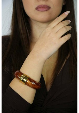 Un bracciale lucido color ambra con superficie liscia e dettagli in metallo color oro, indossato su un polso contro uno sfondo di tessuto scuro.