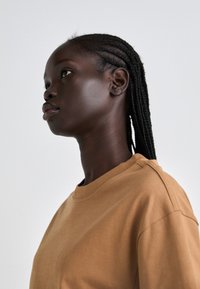 Brun bomulls t-skjorte med rund hals. Modellen har rett, fletter og profilbilde som viser glatt hud og et nøytralt uttrykk.