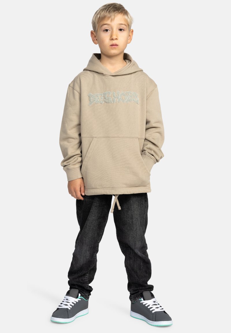 Beige hoodie met een buidelzak en geborduurd logo, gecombineerd met donkerdenim jeans en grijze sneakers met witte veters.