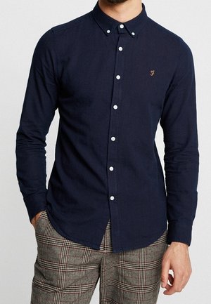 Homme portant une chemise cintrée bleu marine à boutons avec manches longues et un pantalon à carreaux à motifs, debout avec une main dans la poche.