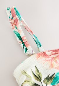 Majica iz tkanine z cvetličnim vzorcem, s roza in zelenimi nastavljivimi naramnicami z volančki. Ima teksturirano belo osnovo z listnatimi poudarki in cvetličnimi detajli.