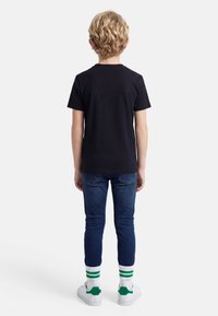 Ξανθό παιδί που στέκεται με την πλάτη, φοράει μαύρο κοντομάνικο μπλουζάκι, μπλε τζιν, άσπρες κάλτσες με πράσινες ρίγες και άσπρα αθλητικά παπούτσια με πράσινες λεπτομέρειες.