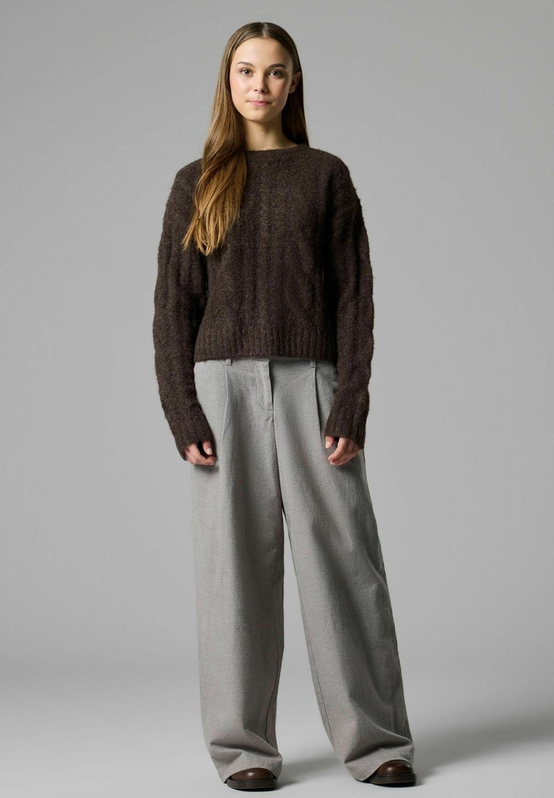 Jeune femme aux longs cheveux bruns porte un pull en tricot marron foncé et un pantalon large gris clair, debout devant un fond gris uni.