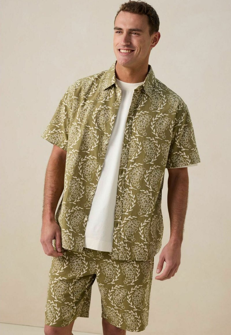 Chemise à manches courtes vert olive et short assorti, tous deux ornés de motifs floraux blancs. La chemise a un col et des boutons ; le short est de longueur genou.