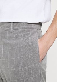 Detailní záběr na osobu oblečenou v šedých kostkovaných kalhotách, s rukou zasunutou v boční kapse, doplněnou o bílou košili.