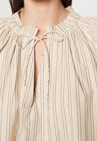Blouse rayée en beige clair avec des lignes beige foncé. Présente un décolleté froncé avec une fermeture à nouer et une coupe légèrement décontractée. Tissu texturé.