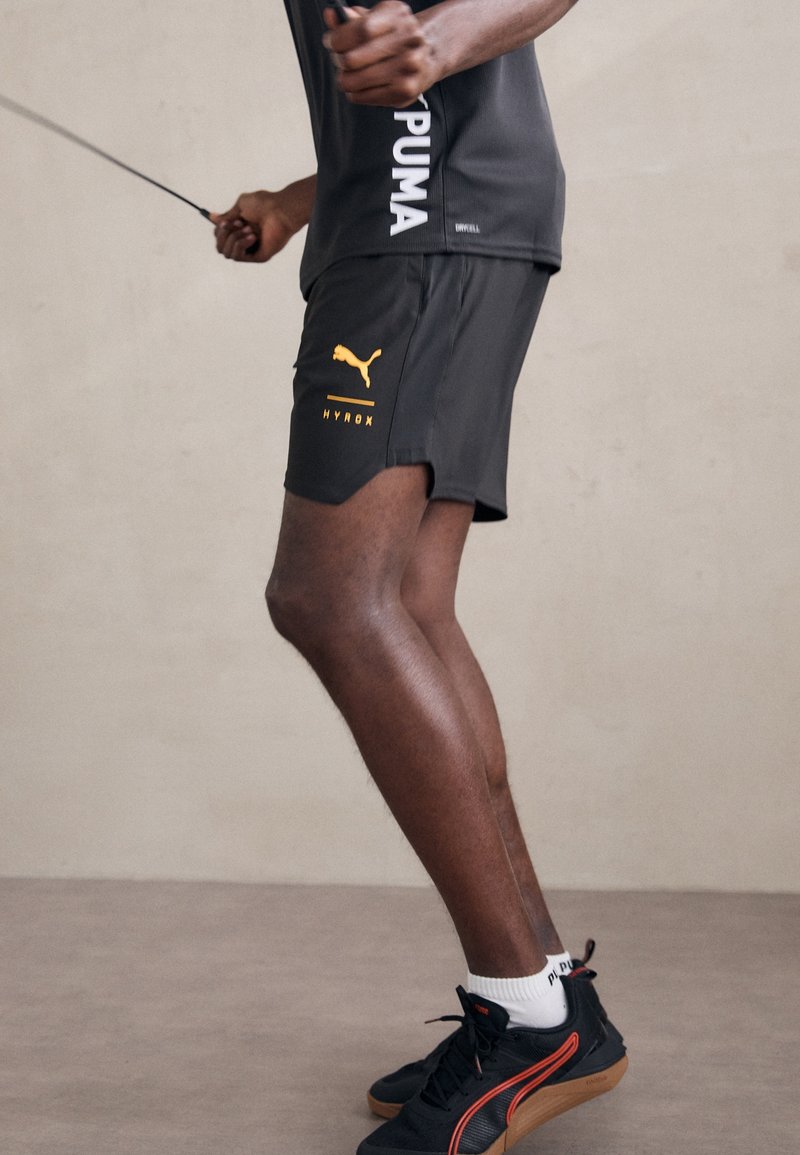 Shorts deportivos negros con el logo amarillo de Puma, combinados con zapatillas negras que tienen acentos rojos. La tela es ligera y texturizada.