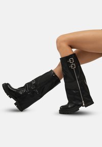 Botas negras hasta la rodilla hechas de un material brillante con una superficie texturizada, que cuentan con un cierre lateral con cremallera y detalles de hebilla. Diseño de suela gruesa.