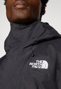 Gros plan d'une personne portant une veste noire avec un col montant, avec le logo "The North Face" brodé sur la poitrine.