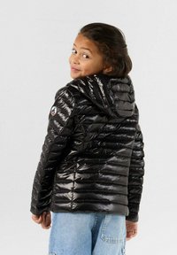 Chaqueta de plumas negra con un acabado brillante, diseño acolchado horizontal, capucha y un parche con logo en el hombro izquierdo. Llevada por un niño.