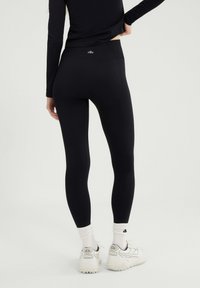 Osoba w czarnych, bezszwowych legginsach, białych skarpetkach sportowych i białych butach do biegania, stojąca na tle jasnego, jednolitego tła.