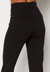 Zwart hoogwaisted broek van gladde stof, voorzien van twee achterzakken en een getailleerde pasvorm met subtiele zijnaaddetails.