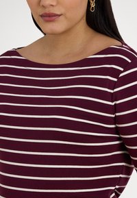 Sieviete, tērpusies bordo un baltā horizontāli svītrainā krekliņā ar zelta ķēdes auskariem, redzama no kakla līdz viduklim.