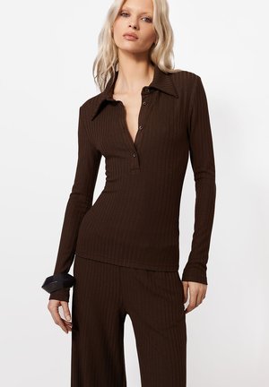 Blondynka ubrana w brązową prążkowaną bluzkę z długim rękawem, kołnierzykiem i guzikami oraz dopasowane spodnie, stojąca na białym tle.