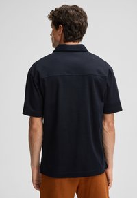 Kurzärmliges, mit Kragen versehenes Hemd aus dunkelblauem Material, mit einer geraden Rückennaht und feiner Textur. Kombiniert mit braunen Hosen.