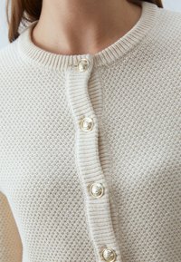 Ljust beige stickad kofta med ett texturerat mönster, ribbad halsringning och guldtonade knappar framtill för stängning.