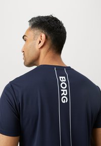 Άνδρας με κοντά σκούρα μαλλιά που φοράει ναυτική μπλε μπλούζα με κάθετο λευκό κείμενο "BORG" στην πίσω πλευρά, σε απλό φόντο.