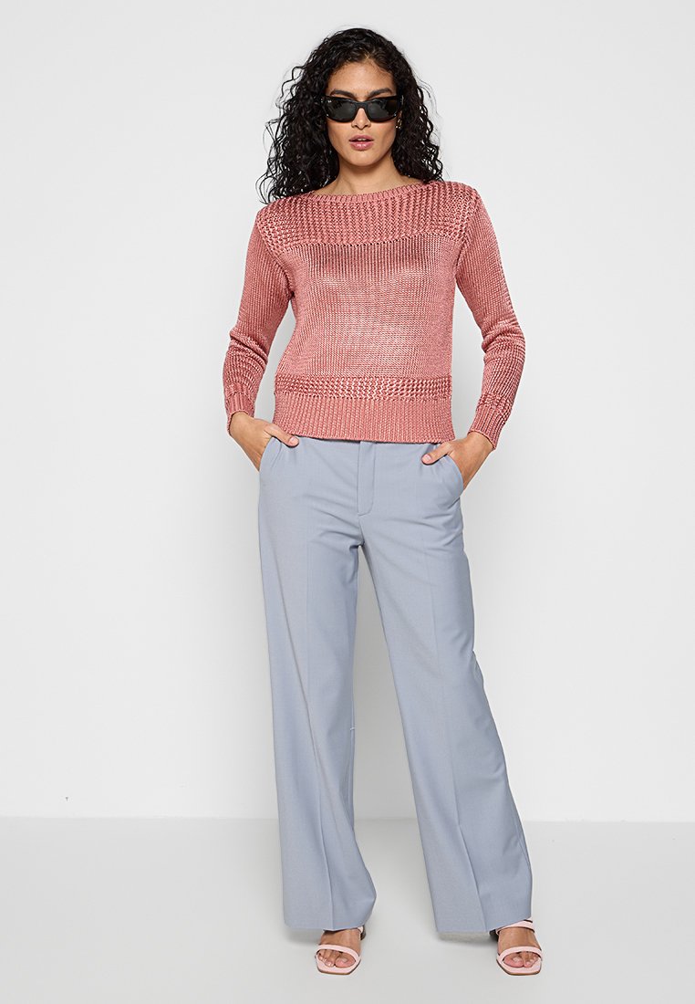 Maglione rosa lavorato a maglia con motivo testurizzato, scollatura rotonda; pantaloni a gamba larga blu chiaro; modella che indossa occhiali da sole, in piedi con le mani in tasca.