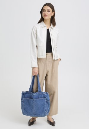 Femeie îmbrăcată în pantaloni bej cu picioare largi și jachetă albă cu dungi, ținând o geantă de umăr din denim albastru, privind ușor spre dreapta cu o expresie neutră.