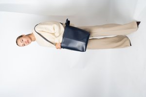 Mulher vestindo um suéter creme e calças largas bege, segurando uma grande bolsa de ombro de couro azul-escuro, de pé contra um fundo branco.