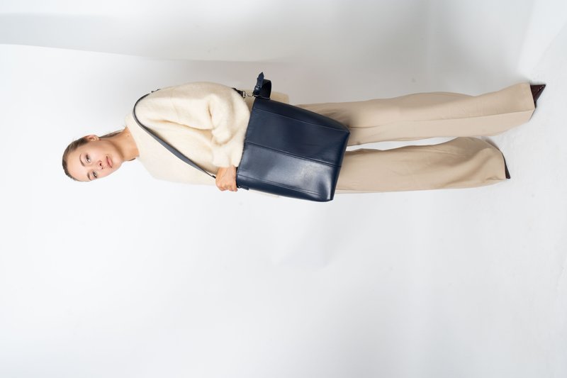 Mujer vestida con un suéter crema y pantalones de pierna ancha beige, sosteniendo un gran bolso de hombro de cuero azul marino, de pie frente a un fondo blanco.