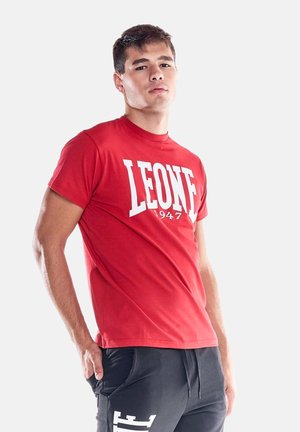 Jonge man die een rood "LEONE 1947" t-shirt en zwarte broek draagt, staat met één hand in zijn zak tegen een effen achtergrond.