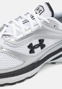 Valkoinen urheilujalkine, jossa on verkkomateriaali, hopeiset yksityiskohdat, musta logo ja valkoiset nauhat. Pohja on musta, jossa on teksturoitu pito ja brändäys.