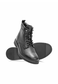 Czarne skórzane botki na sznurówki sięgające kostki z okrągłym noskiem i teksturowaną gumową podeszwą, pokazane z jednym butem stojącym na boku i jednym leżącym na boku.