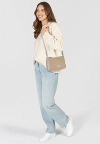 Bolso de mano tejido en beige con acentos en azul claro, forma rectangular, correa ajustable, cierre con cremallera, combinado con un suéter color crema y jeans claros.