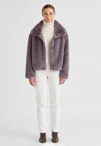 Lila Kunstpelzjacke mit breitem Kragen, vorderen Knöpfen und weicher Textur, getragen über einem cremefarbenen Rollkragenpullover und weißen Hosen.