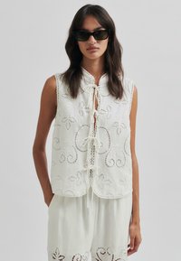 Weißes, besticktes ärmelloses Blusenshirt mit floralen Spitzenverzierungen, hohem Kragen und einer vorderen Schleifenverschlus. Glattes, leichtes Material.