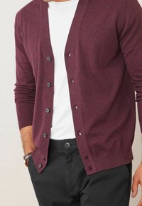 Cardigan bordeaux en tissu doux, avec un col en V, une fermeture boutonnée à l'avant et des poignets côtelés. Porté sur une chemise blanche avec un pantalon noir.