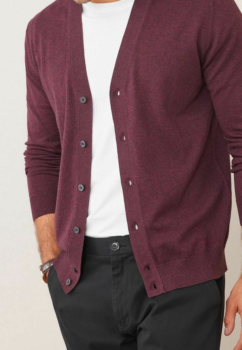 Cardigan bordeaux en tissu doux, avec un col en V, une fermeture boutonnée à l'avant et des poignets côtelés. Porté sur une chemise blanche avec un pantalon noir.