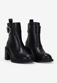 Botas de cuero negras hasta el tobillo con un tacón grueso, cremallera lateral y una banda ancha que presenta un acento circular dorado en el tobillo. Textura suave.