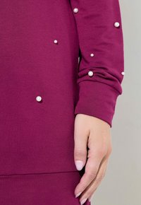 Bordový top s dlouhým rukávem vyrobený z měkké tkaniny, který má dekorativní bílé perly uspořádané asymetricky na rukávech a manžetách.