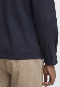 Mörkblå, långärmad skjorta med knäppning vid ärmslut, texturerad tyg, kombinerad med ljusbeige, randiga byxor. Minimalistiska design detaljer synliga.