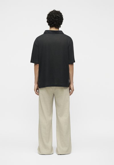 Άτομο με σγουρά μαλλιά που φοράει χαλαρό μαύρο κοντομάνικο πουκάμισο και φαρδιά μπεζ παντελόνια, στέκεται με την πλάτη σε απλό λευκό φόντο.